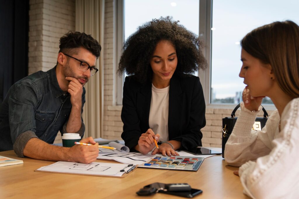 Three professionals collaborate at a table, reviewing design plans and color samples. A coffee cup and a phone are visible, indicating a creative meeting.